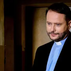Aktor z obsady "Ksiądz Mateusz" w czarnym garniturze i błękitnej koszuli z białym kołnierzykiem, zamyślony.