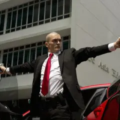 Agent 47 w czarnym garniturze i czerwonym krawacie, z dwoma pistoletami w dłoniach, gotowy do akcji.
