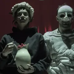 Trzy postacie w maskach na tle czerwonej kurtyny. Jedna z nich trzyma dzbanek z malinami. Scena z "Balladyny" w Teatrze Telewizji.