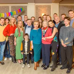 Wspólna obsada serialu "Aktor na wspólnej" pozuje do zdjęcia. Wszyscy uśmiechnięci, w kolorowych strojach, świętują sukces.