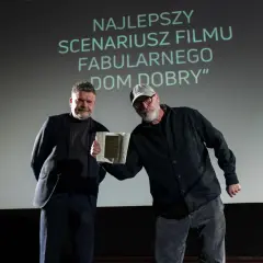 Dwóch mężczyzn na scenie, jeden odbiera nagrodę. Na ekranie napis "NAJLEPSZY SCENARIUSZ".
