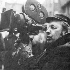Operator filmowy w czapce i płaszczu patrzy przez wizjer kamery filmowej.