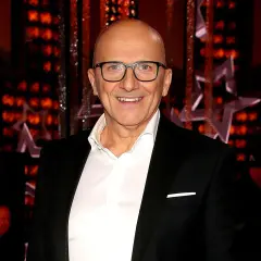 Robert Rozmus w czarnym garniturze i białej koszuli, uśmiecha się na tle czerwonych świateł i gwiazd.