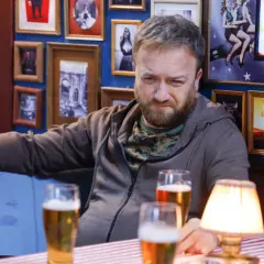 Tatusiowie serial obsada: Mężczyzna z brodą i w bluzie siedzi przy stole z piwem, w tle ściana z ramkami.
