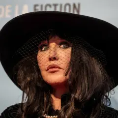 Emma de Caunes w czarnym kapeluszu i welonie, z perłami na szyi, patrzy w górę.