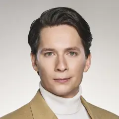 Michał Barczak, aktor, w beżowym płaszczu i białym golfie, z charakterystyczną fryzurą i przenikliwym spojrzeniem.