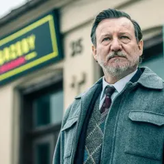 Mężczyzna w eleganckim stroju, z siwą brodą, stoi przed budynkiem z neonem. To część obsady serialu "Minuta ciszy".