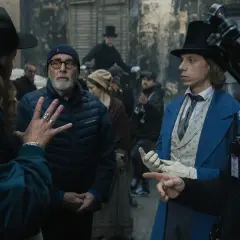 Grupa ludzi na planie filmowym, w tym reżyser w czapce i młody aktor w niebieskim fraku.