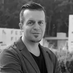 Czarno-białe zdjęcie Marcina Wrony, reżysera, w marynarce, na tle budynków i zieleni.