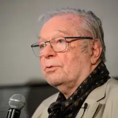 Portret starszego mężczyzny w okularach, mówiącego do mikrofonu, ubranego w jasną marynarkę i wzorzysty szalik.