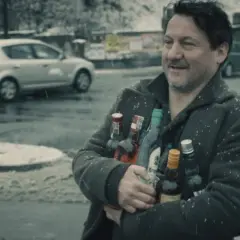 Mężczyzna z uśmiechem niesie kilka butelek alkoholu pod mocnym aniołem obsada. W tle śnieg i samochody.