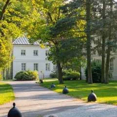 Biały dworek otoczony zielenią, z alejką i ozdobnymi kulami.