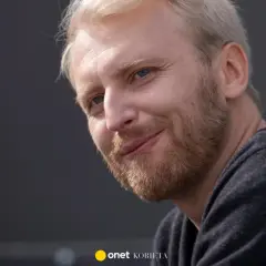 Uśmiechnięty Dobromir Dymecki z jasnymi włosami i niebieskimi oczami, w ciemnej bluzie.