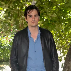 Alain Fabien Delon w czarnej kurtce i niebieskiej koszuli, stoi na tle zieleni.