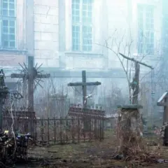 Mglisty cmentarz z kamiennymi krzyżami i zaroślami przed budynkiem opactwa.
