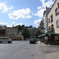 Plac miejski z hotelem, samochodami i kawiarnią pod zielonymi parasolami.