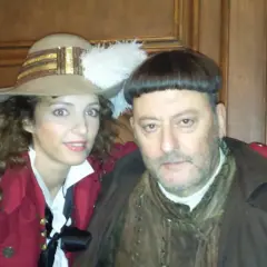 Goście, goście, obsada w historycznych strojach. Kobieta w kapeluszu z piórem i mężczyzna w brązowym płaszczu pozują do zdjęcia.