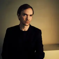 Portret Jana Komasy, reżysera w czarnym garniturze, na tle oświetlonej ściany.