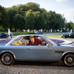 Kobieta w czerwonej sukience wychyla się z okna błękitnego klasycznego samochodu.