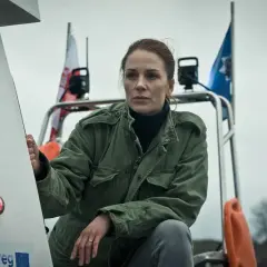 Katarzyna Wajda, aktorka, w zielonej kurtce, z polską flagą w tle, na tle pochmurnego nieba.