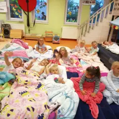Dzieci leżą w śpiworach i kołdrach, gotowe na noc w przedszkolu. Niektóre machają do aparatu, inne patrzą z zaciekawieniem.