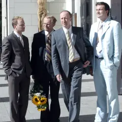 Czterech mężczyzn w garniturach, jeden z bukietem słoneczników, jakby po scenie z filmu "testosteron".