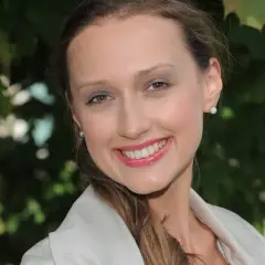 Uśmiechnięta Anna Gzyra w jasnej marynarce, z błyszczącymi kolczykami, na tle zielonych liści.