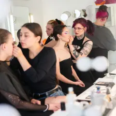 Charakteryzatorka filmowa pracuje nad makijażem modelki. W tle inne osoby przygotowują się do sesji.