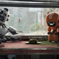 Dwa roboty siedzą przy stole w zniszczonym mieście. Jeden, biały, patrzy na drugiego, pomarańczowego, który pochyla się nad czymś na stole. Scena przypomina "Miłość, śmierć i roboty".