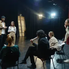 Grupa ludzi na scenie, dyskutująca o sztuce. Zastanawiają się, ile zarabia reżyser, analizując scenariusz.
