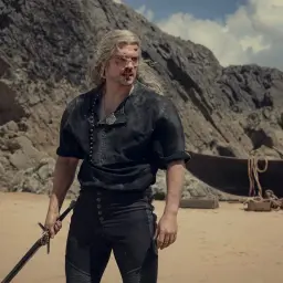 Wiedzmin Geralt z Rivii, ranny i zmęczony, stoi na piaszczystej plaży z mieczem w dłoni. W tle widać łódź i skaliste wybrzeże.
