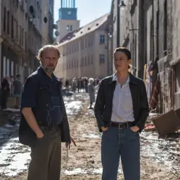 Para zniszczonego miasta po powodzi. Mężczyzna i kobieta stoją na błotnistej ulicy, w tle widać zniszczenia i ludzi. To scena z filmu "Rojst".