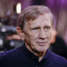 Zbliżenie na twarz Jana Englerta, aktora o niebieskich oczach, w granatowym swetrze. W tle rozmyte światła i sylwetki.