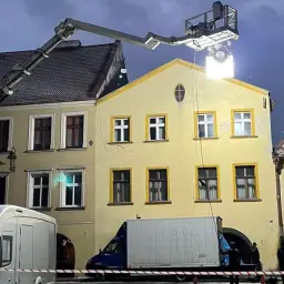 Na planie filmowym, gdzie kręcono "Kolory zła: Czerń", jasny reflektor oświetla żółtą kamienicę. W tle widać ciężarówki i ekipę filmową.