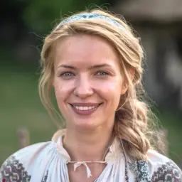 Uśmiechnięta Paulina Gałązka w tradycyjnej, haftowanej koszuli i opasce na włosach, na tle zieleni.