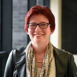 Uśmiechnięta Monika Kurska w okularach i skórzanej kurtce, z apaszką w kratkę i perłowym naszyjnikiem.