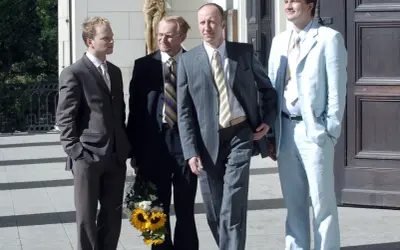 Czterech mężczyzn w garniturach, jeden z bukietem słoneczników, jakby po scenie z filmu "testosteron".
