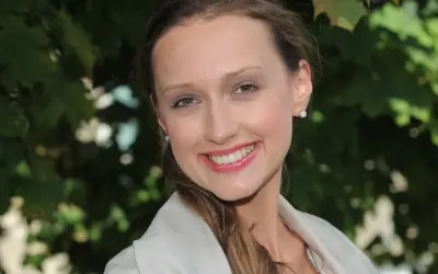Uśmiechnięta Anna Gzyra w jasnej marynarce, z błyszczącymi kolczykami, na tle zielonych liści.