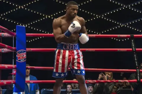 Michael B. Jordan jako Adonis Creed w szortach w barwach USA, gotowy do walki. W tle widać Sylvestra Stallone. Creed II obsada w akcji.