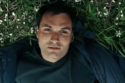 Mężczyzna leży na trawie, otoczony zielenią i białymi kwiatkami. Może to scena z filmu, gdzie wino truskawkowe obsada odgrywa ważną rolę.