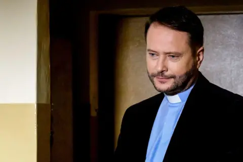 Aktor z obsady "Ksiądz Mateusz" w czarnym garniturze i błękitnej koszuli z białym kołnierzykiem, zamyślony.