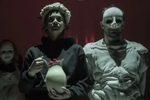 Trzy postacie w maskach na tle czerwonej kurtyny. Jedna z nich trzyma dzbanek z malinami. Scena z "Balladyny" w Teatrze Telewizji.
