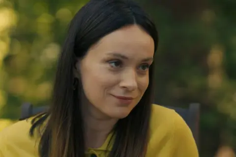 Uśmiechnięta Maria Dejmek w żółtym swetrze, z rozpuszczonymi włosami, na tle zieleni.