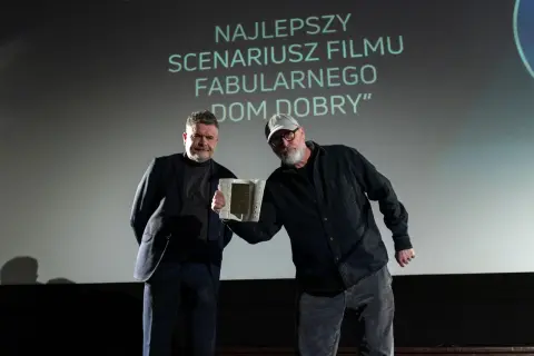 Dwóch mężczyzn na scenie, jeden odbiera nagrodę. Na ekranie napis "NAJLEPSZY SCENARIUSZ".