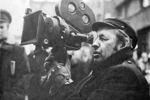 Operator filmowy w czapce i płaszczu patrzy przez wizjer kamery filmowej.