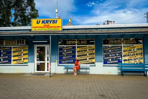 Dziewczyna w czerwonej sukience siedzi na ławce przed sklepem "U Krysi", gdzie można kupić wszystko, od zniczy po narzędzia.