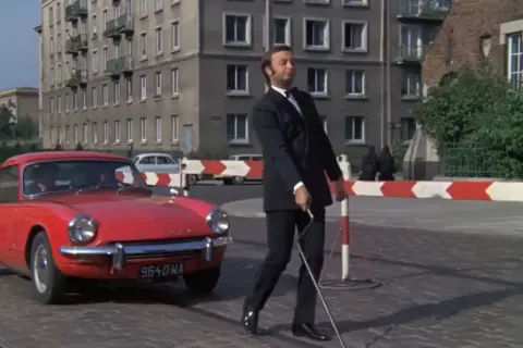 Mężczyzna w garniturze z laską idzie obok czerwonego samochodu. Chyba nie lubię poniedziałku, bo mam ochotę uciec tym autem.