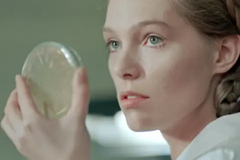 Małgorzata Braunek, młoda kobieta z warkoczem, bada pod mikroskopem szalkę Petriego.