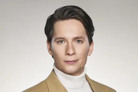 Michał Barczak, aktor, w beżowym płaszczu i białym golfie, z charakterystyczną fryzurą i przenikliwym spojrzeniem.