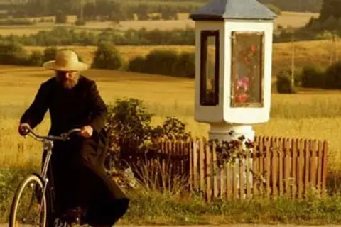 Mężczyzna w czarnym habicie i słomkowym kapeluszu jedzie rowerem obok kapliczki. Czuje się jak u pana boga w ogródku.
